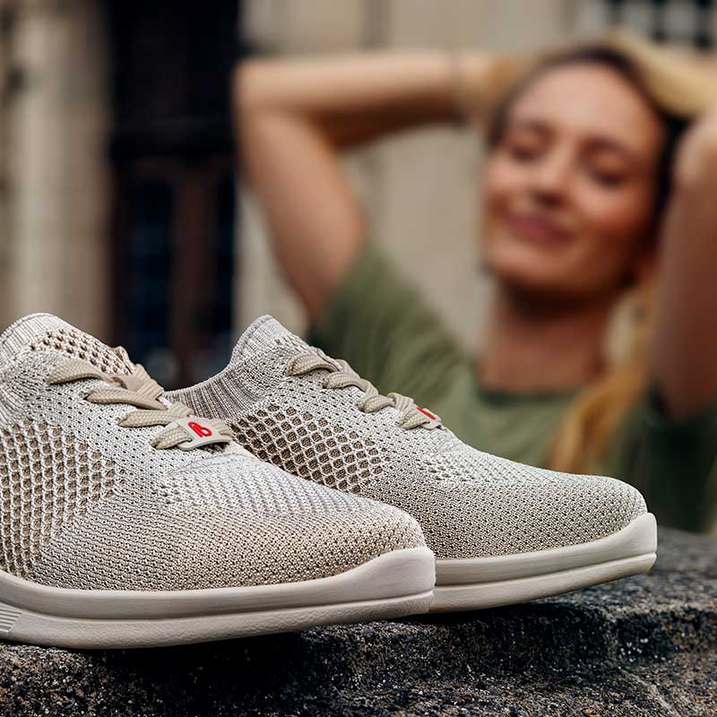 Beigefarbene Berkemann-Sneaker Elea aus 3D AIRKNIT mit hellen Schnürsenkeln, im Hintergrund unscharf eine lächelnde Frau in grünem Shirt.