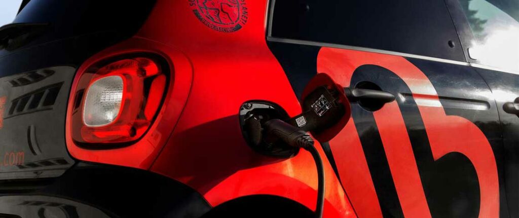 Schwarzer und roter Elektro-Smart mit großem Berkemann-Logo lädt an einer Ladesäule, die Steckverbindung ist im hinteren Fahrzeugbereich eingesteckt.