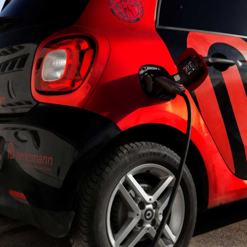 Schwarzer und roter Elektro-Smart mit großem Berkemann-Logo lädt an einer Ladesäule, die Steckverbindung ist im hinteren Fahrzeugbereich eingesteckt.